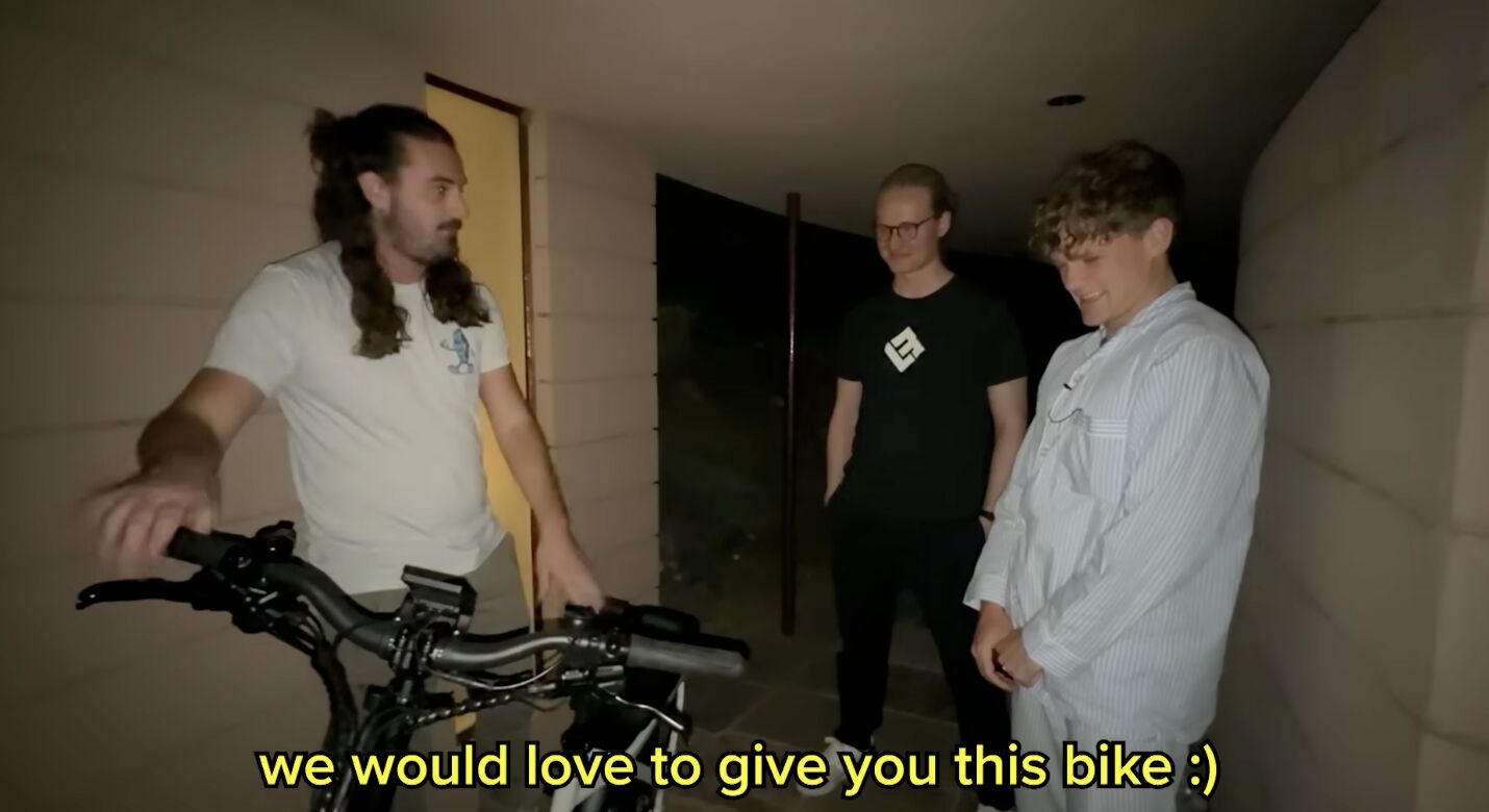 Indoor scene the Lectric team donating a black Lectric bike to Ryan Trahan, who is smiling broadly. Yellow subtitle text reads "we would love to give you this bike :)"