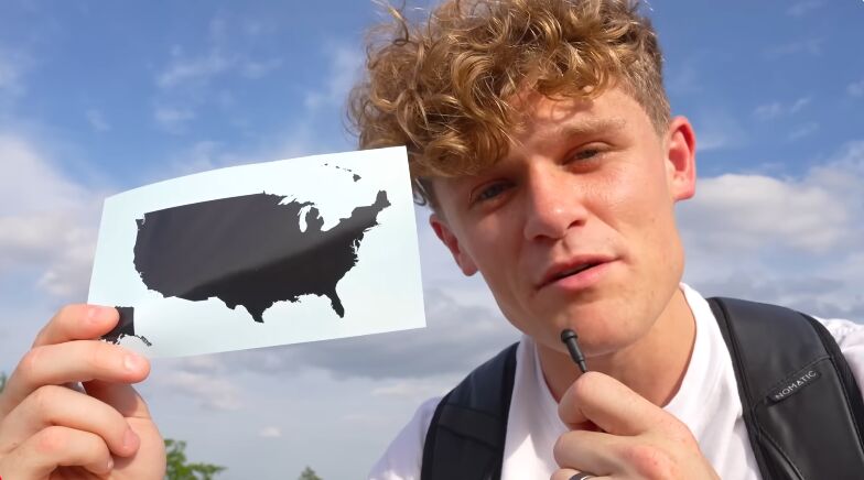 Ryan Trahan outdoors against a blue sky with clouds, holding up a white card or paper showing the silhouette of the United States map. 