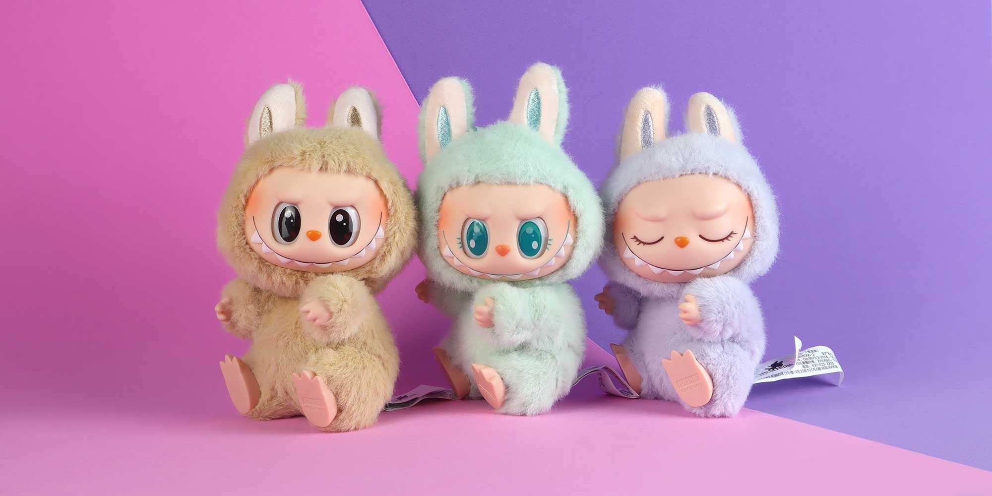 Product photography showing three Labubu plush toy characters with bunny-like ears in pastel colors (beige, mint green, and lavender) against a pink and purple gradient background. 