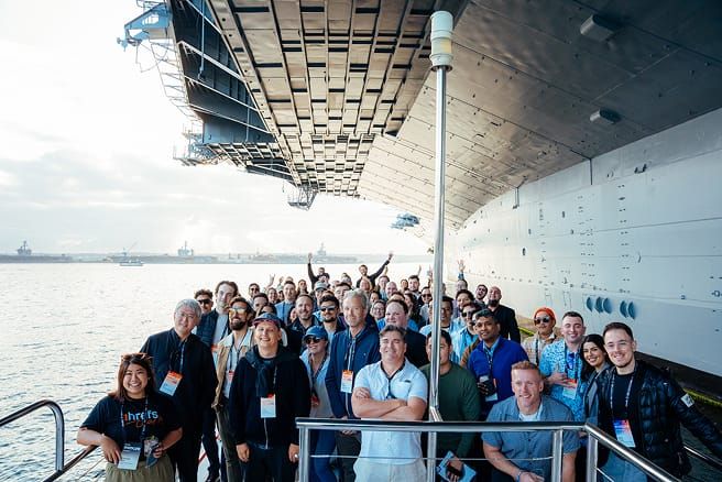 A large group of people smiling and posing for a photo on the deck of a battleship, with water and other ships in the background.
