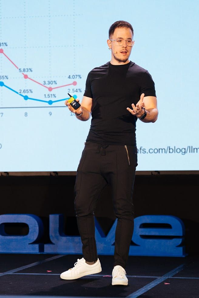 A man in glasses presents on stage, holding a clicker. A line graph with decreasing percentages is projected behind him.