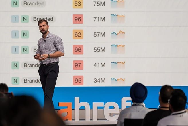 A speaker presents in front of a screen displaying a list of metrics with "Branded" text, numbers, and trend charts.