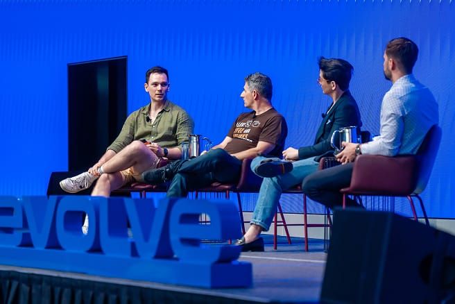 Four men sit on a stage with a blue background, engaged in a panel discussion. "EVOLVE" is on the stage.