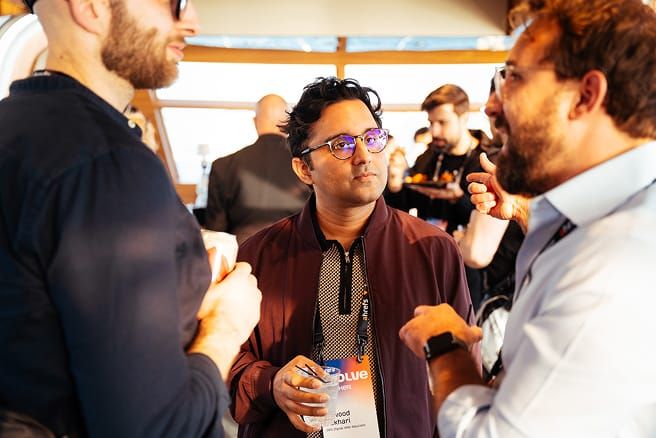 Three men are conversing, two focusing on the man in the center. The man in the center wears glasses and holds a drink.